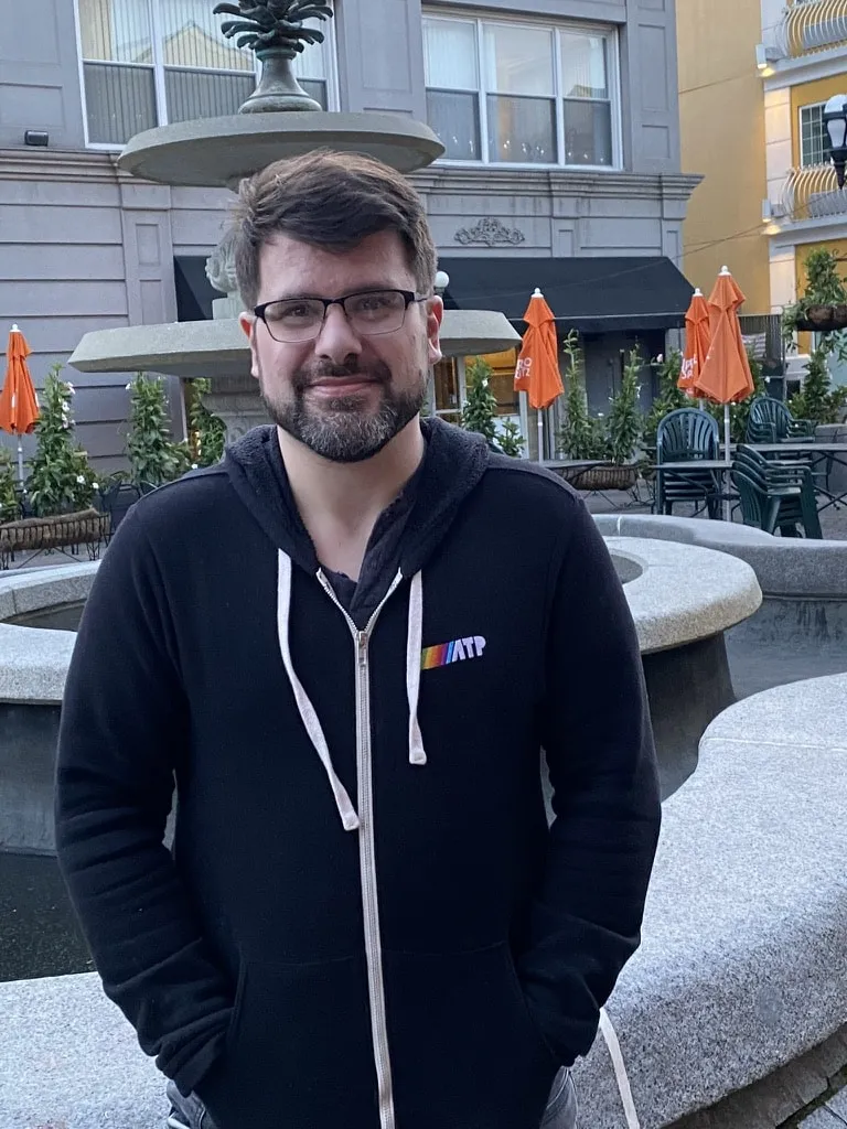 A man with glasses and a beard wearing a black zip-up hoodie stands in front of a decorative fountain in what appears to be an outdoor plaza or courtyard, with buildings and orange umbrellas visible in the background.